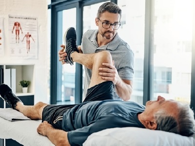 A male physiotherapist helps an older man stretch his leg during a therapy session, with anatomical charts and natural light in the background.