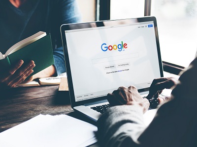 A person typing on a laptop showing the Google UK search page while another person reads a book beside them at a wooden desk.