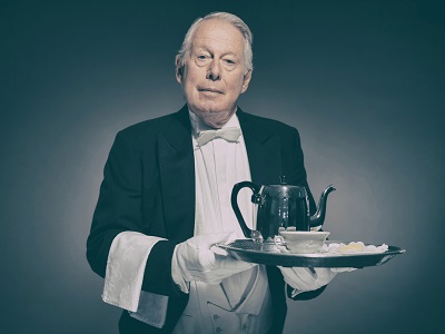 An older man dressed as a traditional British butler holding a silver tray with a teapot, cup, and saucer, standing formally against a plain background.