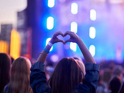 A teenage girl at a live concert forming a heart shape with her hands in front of a brightly lit stage, surrounded by a crowd of people enjoying the music.