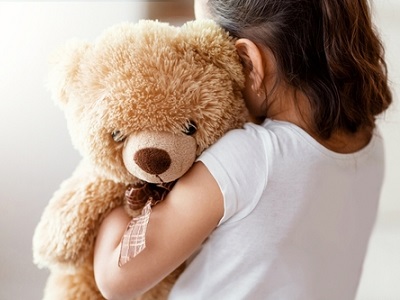 A young child seen from behind hugging a soft brown teddy bear tightly against their shoulder in gentle, natural light.