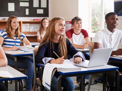A group of teenage students sitting in a bright classroom, smiling and engaged, with notebooks, textbooks, and a laptop on their desks, paying attention to a lesson.