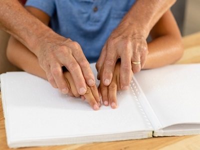 Adult guiding a child’s hands as they read from a Braille book on a wooden table, showing shared touch reading and support for a visually impaired learner.