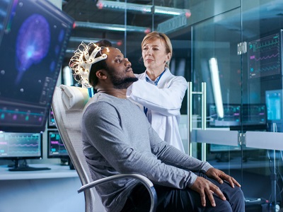 A man wearing an EEG cap sits in a modern lab while a doctor adjusts the headset, with brain scans and wave patterns displayed on large computer screens around them.