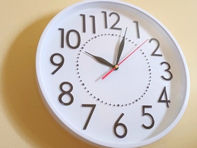 A wall clock with a white face and bold black numbers showing the time as just after 11 o'clock, mounted on a beige wall, symbolising time management or productivity.