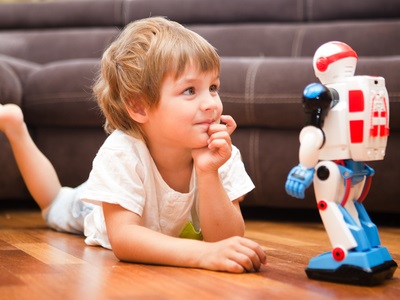 A young child lies on the floor smiling and curiously watching a white and red toy robot standing on the wooden floor, with a sofa in the background.