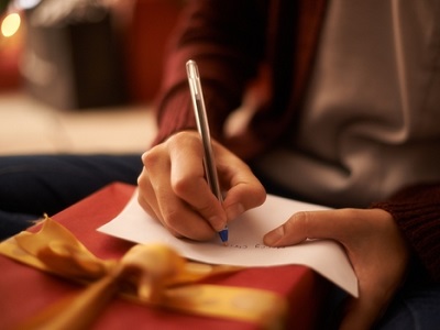 Close-up of a person sitting down and writing a message on white paper resting on a wrapped Christmas present with a golden ribbon.