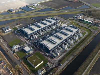 Aerial view of a large industrial data centre surrounded by green fields, canals, and roads, showing cooling units, infrastructure buildings, and nearby farmland.