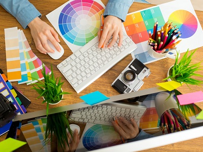 A person using a computer at a colourful creative workstation, surrounded by colour swatches, a keyboard, pencils, a camera, and small plants — representing graphic design or visual creativity.