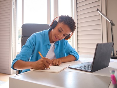 A teenager wearing headphones sits at a tidy desk, smiling while writing in a notebook with a laptop open in front of them, working on schoolwork in a bright, organised bedroom.