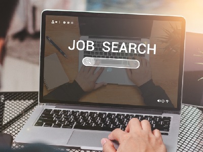 A person using a laptop with the words 'Job Search' on the screen, reflecting hands typing on the keyboard, with plants and a coffee cup on the desk.