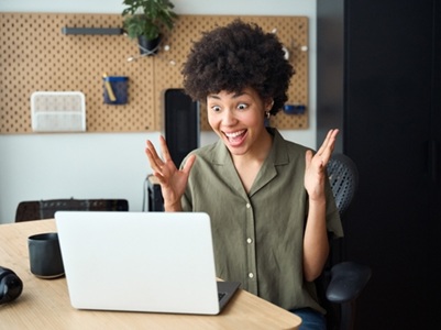 A teenage girl smiling and raising her hands in surprise while looking at a laptop screen, seated at a desk in a modern home or study environment.