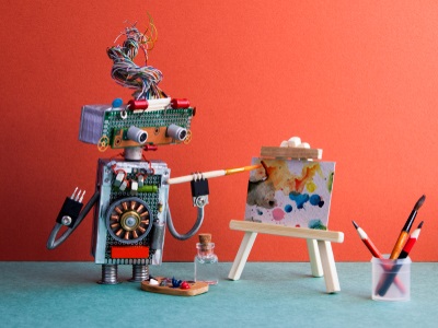 A small, homemade robot holding a paintbrush and painting on a mini canvas, with art supplies and pencils nearby, set against a red and teal background.