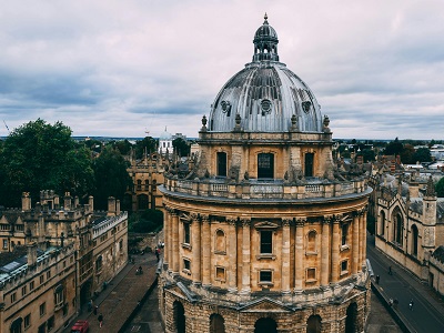 Aerial view of the Radcliffe Camera in Oxford, England, with its large domed roof surrounded by historic university buildings under a cloudy sky.