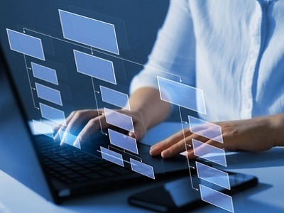 Person typing on a laptop with floating transparent flowchart boxes showing a digital diagram interface, smartphone beside the keyboard, cool blue lighting.