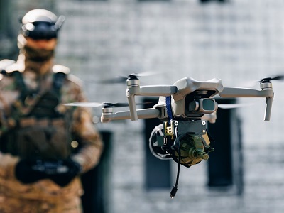A close-up of a military-grade drone carrying equipment, with a soldier in camouflage gear blurred in the background, representing AI use in defence operations.