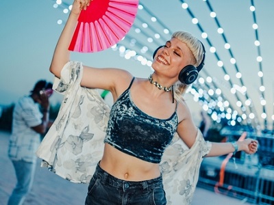A smiling young woman dancing outdoors in the evening, wearing headphones and holding a bright pink fan, with strings of lights glowing in the background.