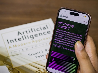A person holding a smartphone showing the OpenAI ChatGPT introduction page, with an artificial intelligence textbook titled “Artificial Intelligence: A Modern Approach” in the background.