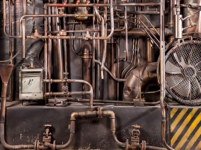 A dense network of old industrial metal pipes, valves and a large fan set against a worn machine wall.
