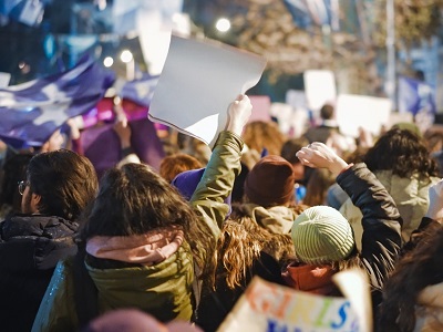 Night-time street protest with a crowd holding placards and flags, people in winter coats and hats, arms raised while marching through a city.