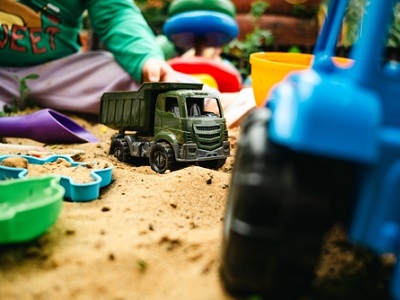 Close-up of a child playing in a sandpit with toy dump trucks, buckets, and moulds; one green truck in focus and a blue truck blurred in the foreground.