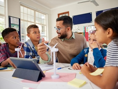 Teacher sitting at a classroom table with a mixed group of children, explaining a science activity that uses small model wind turbines and a digital tablet, as the pupils listen and discuss their ideas together in a bright, modern classroom.