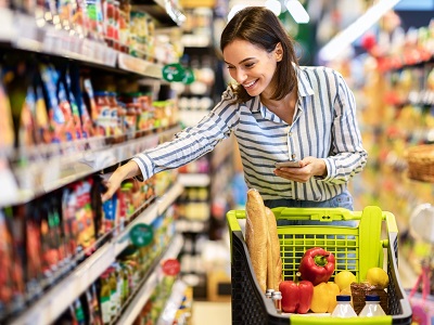 A smiling woman shopping in a supermarket aisle with a trolley full of groceries, symbolising Asda’s trial of AI-powered cameras that scan shelves to detect empty spaces and expired products.