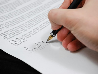 A person signing a formal document with a black fountain pen, close-up of the hand and signature on the page.