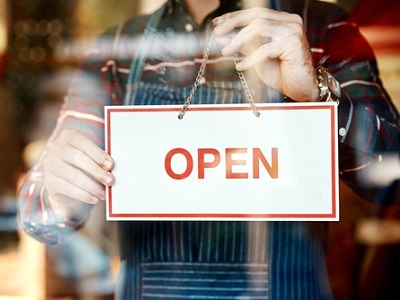 A shopkeeper in an apron holds an “Open” sign on a glass door, representing small businesses receiving new AI-powered support to stay efficient and welcoming.