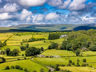 A bright rural South Wales landscape with a patchwork of green fields, hedgerows, scattered farmhouses and woodland, leading to low hills under a blue sky filled with white clouds.