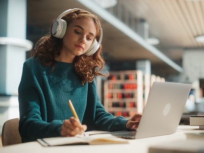 A teenage girl wearing headphones sits at a desk in a modern library, writing in a notebook while using a laptop, focused and studying with books and pens nearby.