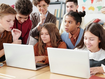 Group of school pupils using laptops at a classroom table while a teacher watches and supports their work.