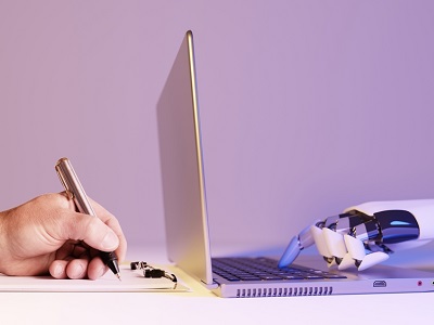 A close-up image showing a human hand writing on paper next to a robotic hand typing on a laptop keyboard, symbolising teamwork and collaboration between humans and artificial intelligence.