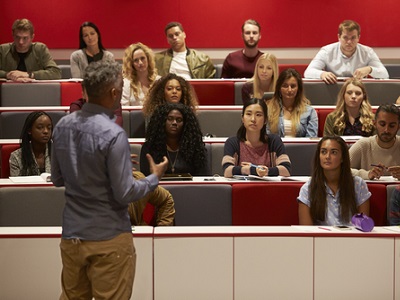 A lecturer speaking to university students seated in a tiered lecture theatre, with students listening and taking notes.
