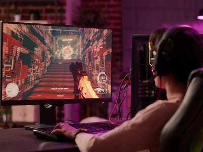 A teenager wearing headphones plays a first-person shooter video game on a computer monitor, representing xAI hiring human tutors to help train its AI, Grok, in game design.
