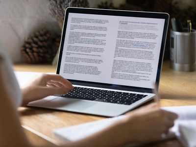 A student sitting at a desk using a laptop to work on a typed essay, with focused hands on the keyboard and notes nearby, in a home or study setting.