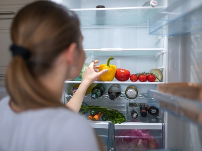 A woman reaching for a yellow bell pepper in a fridge containing fresh vegetables, drinks, and sauces, viewed from behind the open fridge door in a modern kitchen.