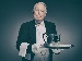 An older man dressed as a traditional British butler holding a silver tray with a teapot, cup, and saucer, standing formally against a plain background.