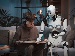 A teenage boy sits on a sofa reading a book while a humanoid robot points to the pages, appearing to help him study in a modern, softly lit room.