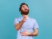A young man with a beard wearing a light blue t-shirt laughs hard, pointing forward with one hand and holding his stomach with the other, against a blue background.