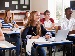 A group of teenage students sitting in a bright classroom, smiling and engaged, with notebooks, textbooks, and a laptop on their desks, paying attention to a lesson.