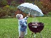 A young child in a raincoat holding an umbrella over a barbecue in the garden while it rains, showing food cooking on the grill despite the wet weather.