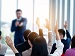 A group of business professionals raising their hands during a seminar or workshop, with a presenter speaking at the front of a bright, modern conference room.