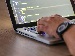 Close-up of a person’s hands typing on a laptop keyboard with computer code displayed on the screen, symbolising programming and AI coding work.