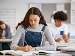 Teenage girl sitting at a classroom desk, concentrating as she writes in an exercise book with open textbooks around her, while other students work in the background, suggesting a quiet exam or homework-style study session.