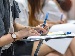 Close-up of students writing in an exam-style classroom, using pen and paper on chair-mounted desks, with rows of seats and a soft background blur.