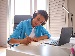 A teenager wearing headphones sits at a tidy desk, smiling while writing in a notebook with a laptop open in front of them, working on schoolwork in a bright, organised bedroom.