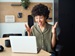 A teenage girl smiling and raising her hands in surprise while looking at a laptop screen, seated at a desk in a modern home or study environment.
