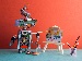A small, homemade robot holding a paintbrush and painting on a mini canvas, with art supplies and pencils nearby, set against a red and teal background.