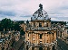 Aerial view of the Radcliffe Camera in Oxford, England, with its large domed roof surrounded by historic university buildings under a cloudy sky.